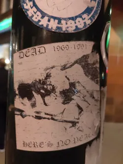 A black and white sticker with an image and text. The text says "Dead 1969-1991" at the top and "Here's No Peace" at the bottom. The image is a blurry depiction of what seems like a person or a group of people. The sticker is slightly damaged and has a worn appearance.