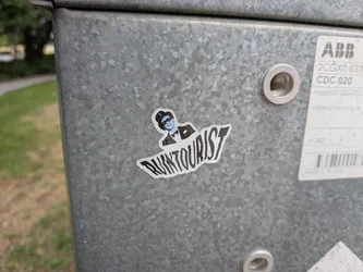 Street sticker Die-cut sticker depicting a blue-skinned character wearing a black top hat, domino mask, and tuxedo. Below the character, the stylized text 'RUINTOURIST' is printed in bold black font. The sticker has a white outline and is affixed to a galvanized metal surface.