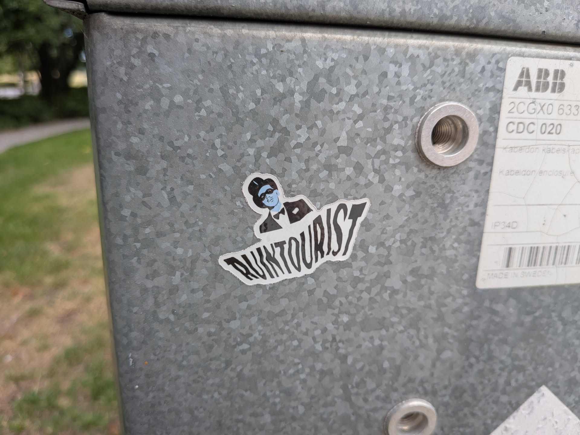 Die-cut sticker depicting a blue-skinned character wearing a black top hat, domino mask, and tuxedo. Below the character, the stylized text 'RUINTOURIST' is printed in bold black font. The sticker has a white outline and is affixed to a galvanized metal surface.