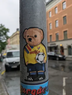 Street sticker This sticker features a brown teddy bear wearing a yellow soccer jersey with blue and red stripes on the shoulder, the word "mita" printed on the front, and black Adidas shorts and shoes. It is holding a soccer ball. The bottom of the pole also has another sticker which reads "Djurgård"