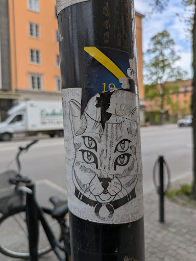 A black and white sticker of a cat's head with four eyes, wearing a collar with a ring. The sticker is applied to a black pole.
