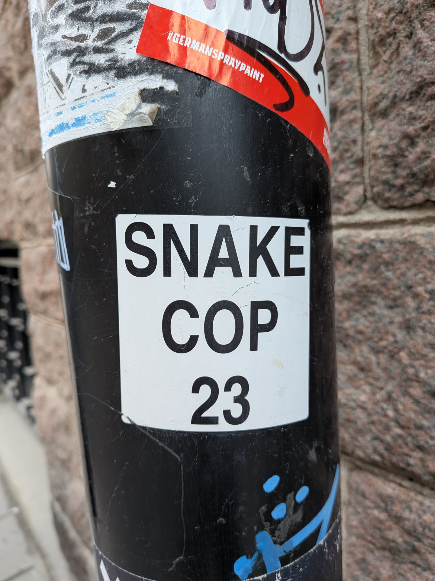 The central sticker is a rectangular white label featuring bold, black sans-serif text. The text is arranged in three lines: 'SNAKE' on the top line, 'COP' on the middle line, and '23' on the bottom line. The sticker is applied to a textured black pole and shows signs of wear, including small cracks in its white surface and slight peeling at the edges, confirming its physical presence and exposure to the elements. The design is minimalist, focusing solely on the impactful typography.