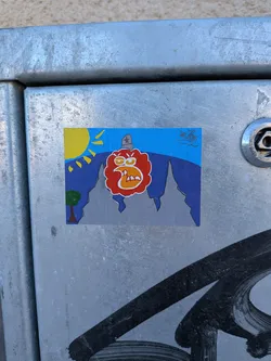 Street sticker A rectangular sticker depicting a stylized landscape with a prominent, angry-looking character. The upper portion features a bright blue sky with a yellow sun and radiating rays in the top left. Below the sky, jagged dark blue mountains transition into grey peaks in the lower half. A small green tree with a brown trunk is visible on the far left. The central figure is a large, red, cloud-like or blob-like creature with an orange outline, featuring angry white eyebrows, yellow eyes with red pupils, and a wide-open mouth revealing jagged white teeth and an orange tongue. It wears a grey, military-style hat with a black star on its head. The overall style is cartoonish and uses vibrant colors.