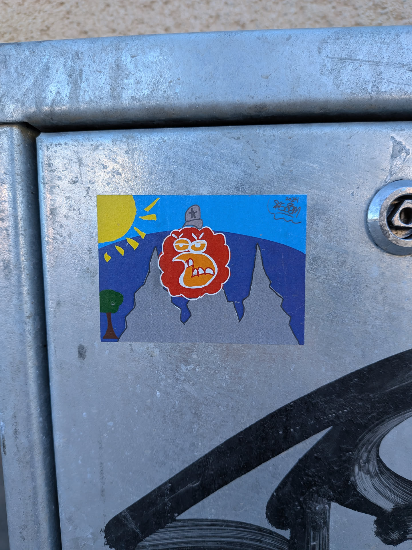 A rectangular sticker depicting a stylized landscape with a prominent, angry-looking character. The upper portion features a bright blue sky with a yellow sun and radiating rays in the top left. Below the sky, jagged dark blue mountains transition into grey peaks in the lower half. A small green tree with a brown trunk is visible on the far left. The central figure is a large, red, cloud-like or blob-like creature with an orange outline, featuring angry white eyebrows, yellow eyes with red pupils, and a wide-open mouth revealing jagged white teeth and an orange tongue. It wears a grey, military-style hat with a black star on its head. The overall style is cartoonish and uses vibrant colors.