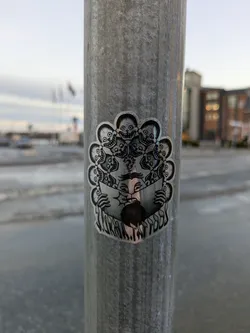 A silver holographic sticker featuring a central figure reading a book while being encircled by numerous skull faces. The style is dark, slightly macabre, yet artistic. The sticker is affixed to a metal pole outdoors.