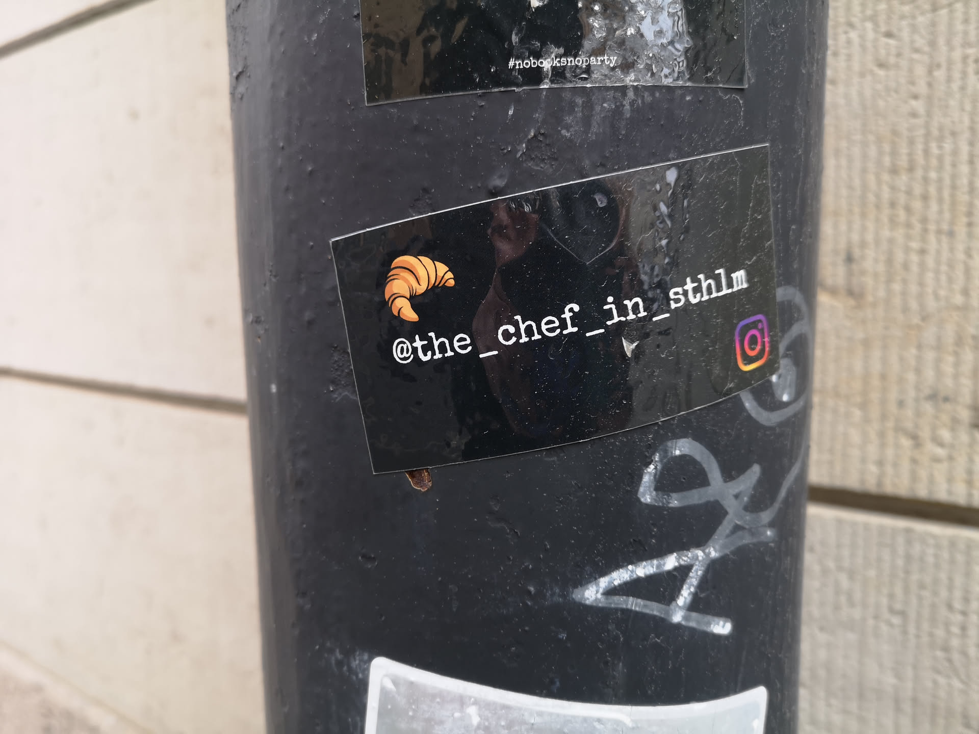 The sticker is rectangular with a glossy black background. On the left side, there is a vibrant orange and yellow illustration of a croissant. To the right of the croissant, white text reads '@the_chef_in_sthlm'. Further to the right, a colorful Instagram logo, displaying its characteristic gradient of orange, pink, and purple, is visible. The sticker has a reflective surface and appears to be adhered to a textured black pole.