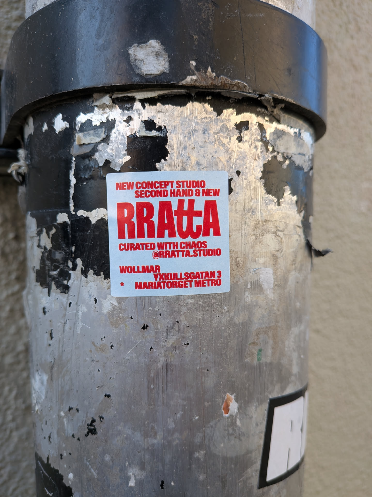 A rectangular white sticker with bold red text, featuring the prominent 'RRATTA' logo in a stylized sans-serif font. Above it, smaller text announces 'NEW CONCEPT STUDIO' and 'SECOND HAND & NEW'. Below the main logo, 'CURATED WITH CHAOS' and the social media handle '@RRATTA.STUDIO' are displayed. The bottom section includes a small red star followed by the address 'WOLLMAR YXKULLSGATAN 3 MARIATORGET METRO'. The sticker is affixed to a weathered metal pole, showing some peeling and wear around its edges.