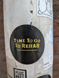 Street sticker A circular sticker with a black background featuring prominent text in a distressed, yellow-gold font. The main phrase reads 'TIME To Go To REHAB', with the word 'REHAB' having a subtle green-yellow gradient. Below this, in smaller white text, is the website 'WWW.THEHOUSEREHAB.COM'. The sticker has a slight holographic or iridescent sheen visible around its edge where it meets the pole, and its surface appears slightly textured or dusty.