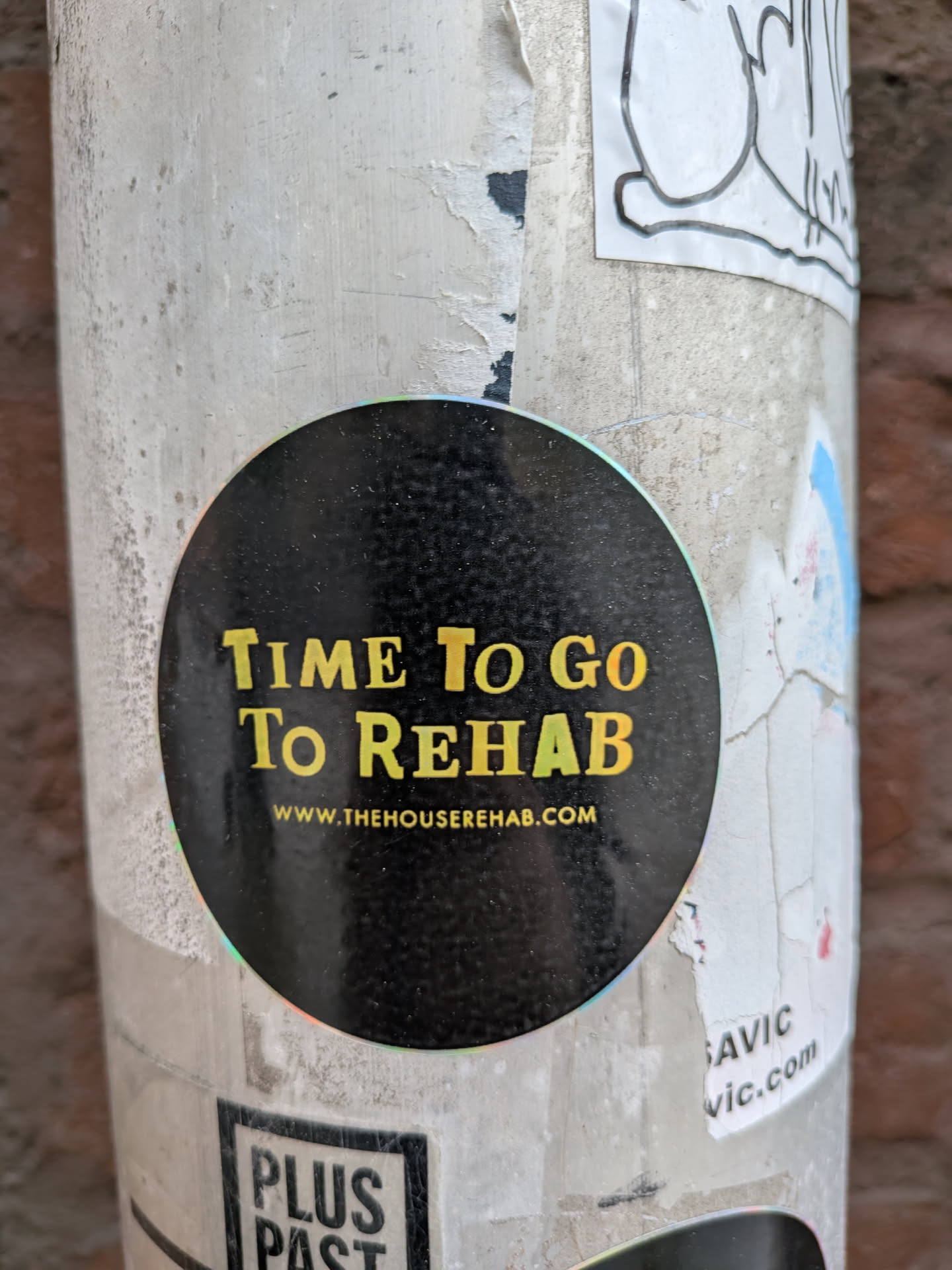 A circular sticker with a black background featuring prominent text in a distressed, yellow-gold font. The main phrase reads 'TIME To Go To REHAB', with the word 'REHAB' having a subtle green-yellow gradient. Below this, in smaller white text, is the website 'WWW.THEHOUSEREHAB.COM'. The sticker has a slight holographic or iridescent sheen visible around its edge where it meets the pole, and its surface appears slightly textured or dusty.