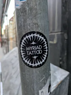 Street sticker This is a black and white circular sticker adhered to a gray metal pole. The sticker features the words "MYRIAD TATTOO" in the center, surrounded by a sunburst-like design composed of white teardrop shapes pointing inwards.