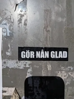 A rectangular black sticker featuring bold, white, sans-serif Swedish text that reads 'GÖR NÅN GLAD'. It is affixed to a weathered grey metallic surface.
