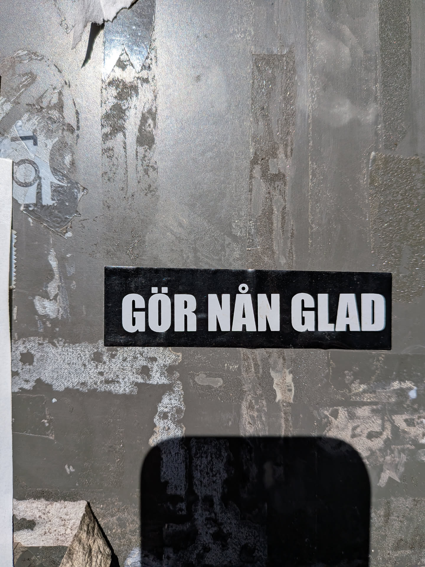A rectangular black sticker featuring bold, white, sans-serif Swedish text that reads 'GÖR NÅN GLAD'. It is affixed to a weathered grey metallic surface.