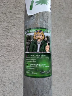 Street sticker The sticker is designed to resemble a Pokemon trading card. It features a cartoonish illustration of a man named Kenta, who has blond hair, sunglasses, a mustache, and is wearing a green jacket over a white sweater. He is smoking a cigarette and giving the middle finger. The background of the illustration is a city street scene with buildings and trees. The card has green borders, text in Swedish, and includes elements like 'LEGEND', 'HP 69', ability descriptions, and stat categories ('Styrka' and 'Svaghet'). A logo reading 'Vintergatan' is visible at the bottom right.