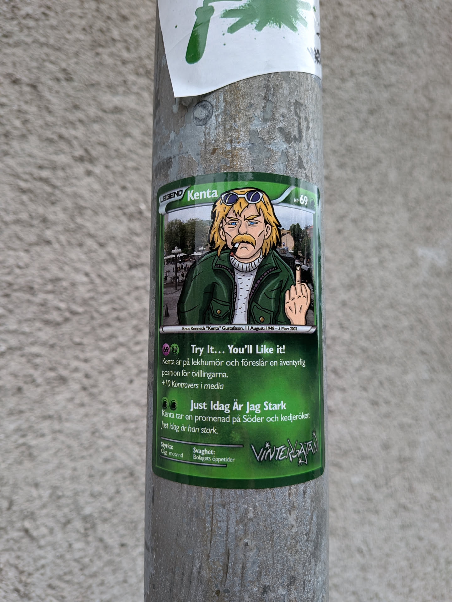 Street sticker The sticker is designed to resemble a Pokemon trading card. It features a cartoonish illustration of a man named Kenta, who has blond hair, sunglasses, a mustache, and is wearing a green jacket over a white sweater. He is smoking a cigarette and giving the middle finger. The background of the illustration is a city street scene with buildings and trees. The card has green borders, text in Swedish, and includes elements like 'LEGEND', 'HP 69', ability descriptions, and stat categories ('Styrka' and 'Svaghet'). A logo reading 'Vintergatan' is visible at the bottom right.