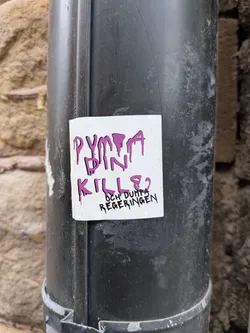 Street sticker A square white sticker with rounded corners is stuck to a black metal pipe. The sticker features stylized, dripping text. The words 'DUMPA DIN KILLE?' are written in a large, dripping purple font, while the phrase 'OCH DUMPA REGERINGEN' is written below in a smaller, dripping black font.