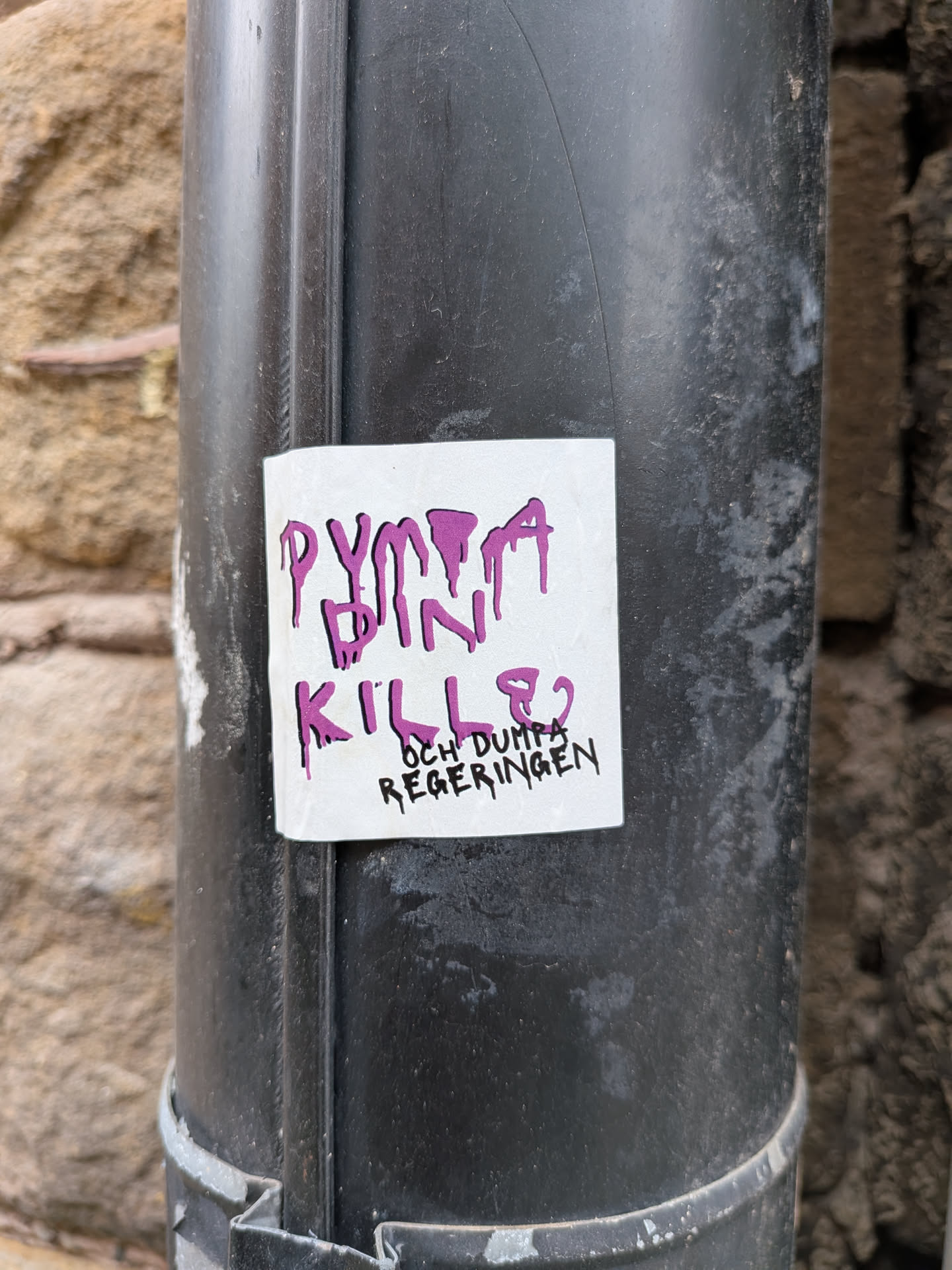 A square white sticker with rounded corners is stuck to a black metal pipe. The sticker features stylized, dripping text. The words 'DUMPA DIN KILLE?' are written in a large, dripping purple font, while the phrase 'OCH DUMPA REGERINGEN' is written below in a smaller, dripping black font.