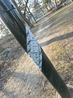 Street sticker The sticker is an elongated, vertically oriented design wrapped around a dark green pole. Its primary background is white, overlaid with dense, intricate black geometric patterns that evoke a sense of urban architecture, circuit boards, or abstract cityscapes. Dominating the center is the word 'CHOPPER', rendered in a bold, blocky, and highly stylized white font with a distinctive light teal or mint green outline and shadow effect, making the text pop against the monochrome background. Below the text, a circular motif, possibly a stylized wheel or gear, is integrated into the geometric framework. The sticker shows minor wear and tear, confirming its physical presence on the pole.