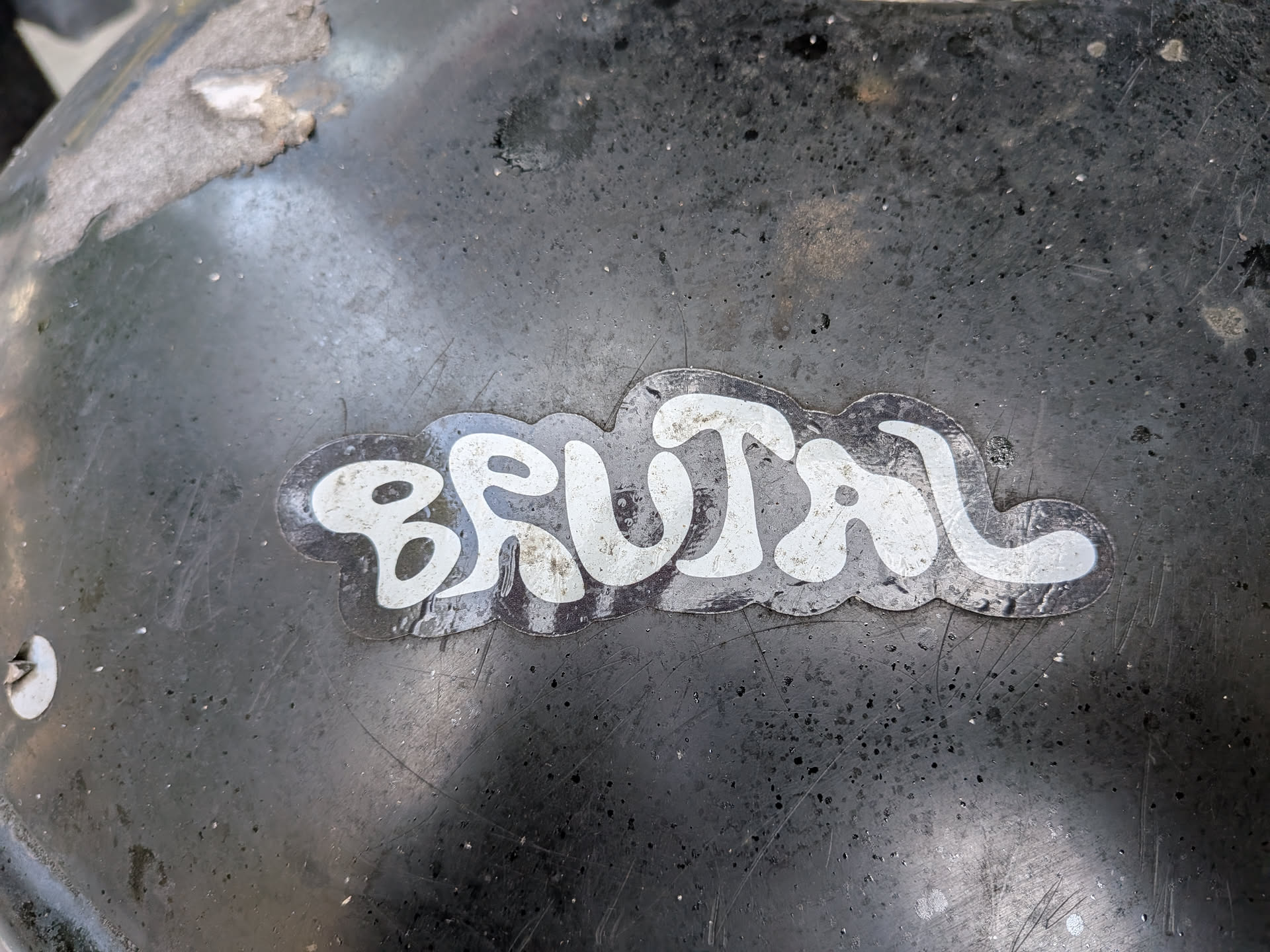 The sticker is centrally located and features the word "BRUTAL" in a stylized, bold, white font with a black outline. The lettering has a slightly distressed, hand-drawn quality. The sticker has a clear, possibly glossy finish and a jagged, irregular edge. It appears to be adhered to a dark, textured surface that shows signs of wear and tear, including scratches and small specks of dirt or debris.