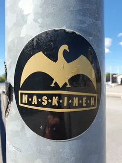 Street sticker A round black and gold sticker featuring a stylized bird design and the text 'M*A*S*K*I*N*E*N'. The sticker is affixed to a metallic pole outdoors.
