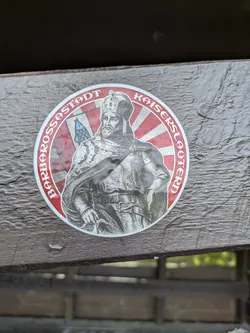 A round sticker features a depiction of Kaiser Barbarossa, the Holy Roman Emperor, against a red and white striped background. The sticker is placed on a wooden surface. Text around the image reads 'Barbarossastadt Kaiserslautern'.