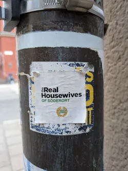 Street sticker The sticker is white with black and green text. The top line reads 'the Real' in a serif font, with 'Real' being larger and bolder. Below that, in a sans-serif font, is 'Housewives'. The bottom line reads 'OF SÖDERORT' in green, also in a sans-serif font. In the lower center of the sticker is a circular emblem with a green and white design, possibly a coat of arms, surrounded by a gold wreath. The sticker is slightly creased and appears to be adhered to a dark, weathered pole, with a blue and yellow patterned layer underneath. There are also some blue and yellow paint splatters on the surface of the pole around the sticker.