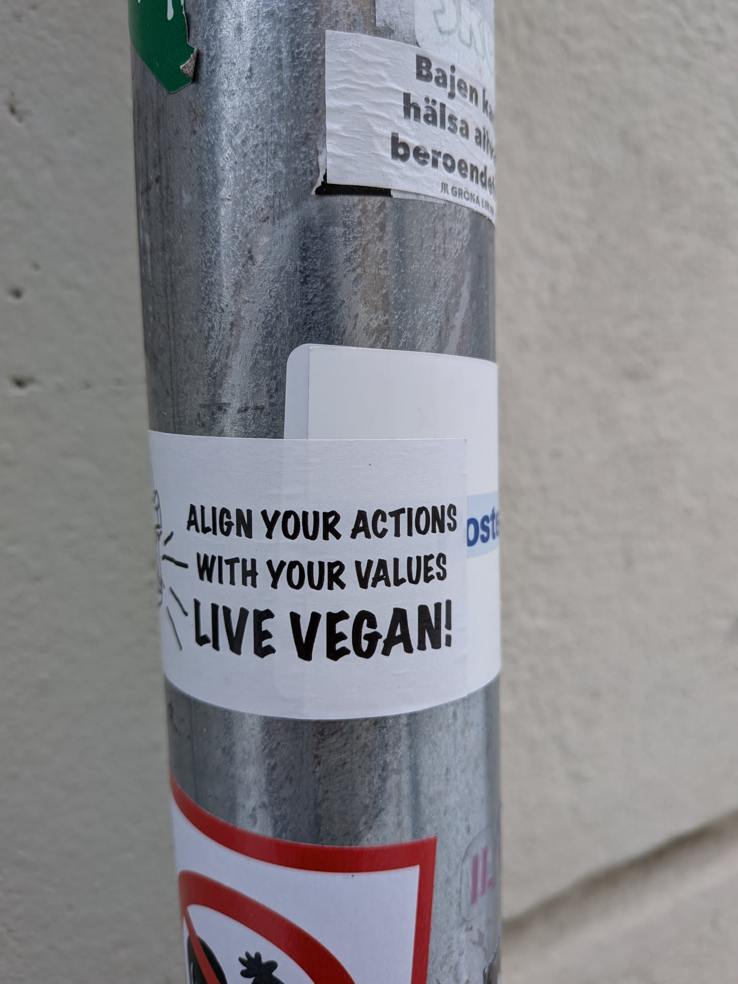 Street sticker A rectangular white sticker with bold black text, adhered to a textured metallic pole. The text is arranged in three lines: 'ALIGN YOUR ACTIONS', 'WITH YOUR VALUES', and 'LIVE VEGAN!'. To the left of the first line, there is a simple black line drawing resembling a head with radiating lines, possibly symbolizing thought or communication. The sticker shows minor wrinkles and texture, suggesting it is a physical sticker.