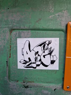 A square sticker with a black and white graffiti-style design. The graffiti appears to say "AGZ". The sticker is affixed to a weathered, green surface.