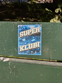 Street sticker The sticker is predominantly blue and white, featuring a dense crowd of people with their arms raised, many holding flags. The word 'SUPER' is emblazoned in large, bold, white, 3D-effect letters across the top half of the sticker, with a blue background behind it. Below 'SUPER', the word 'KLUBL' is similarly presented in large, bold, white, 3D-effect letters. The background behind the crowd appears to be a stylized representation of a stadium or a large gathering, with a blue and white color scheme. The edges of the sticker show some wear and tear, indicating it has been applied for some time.