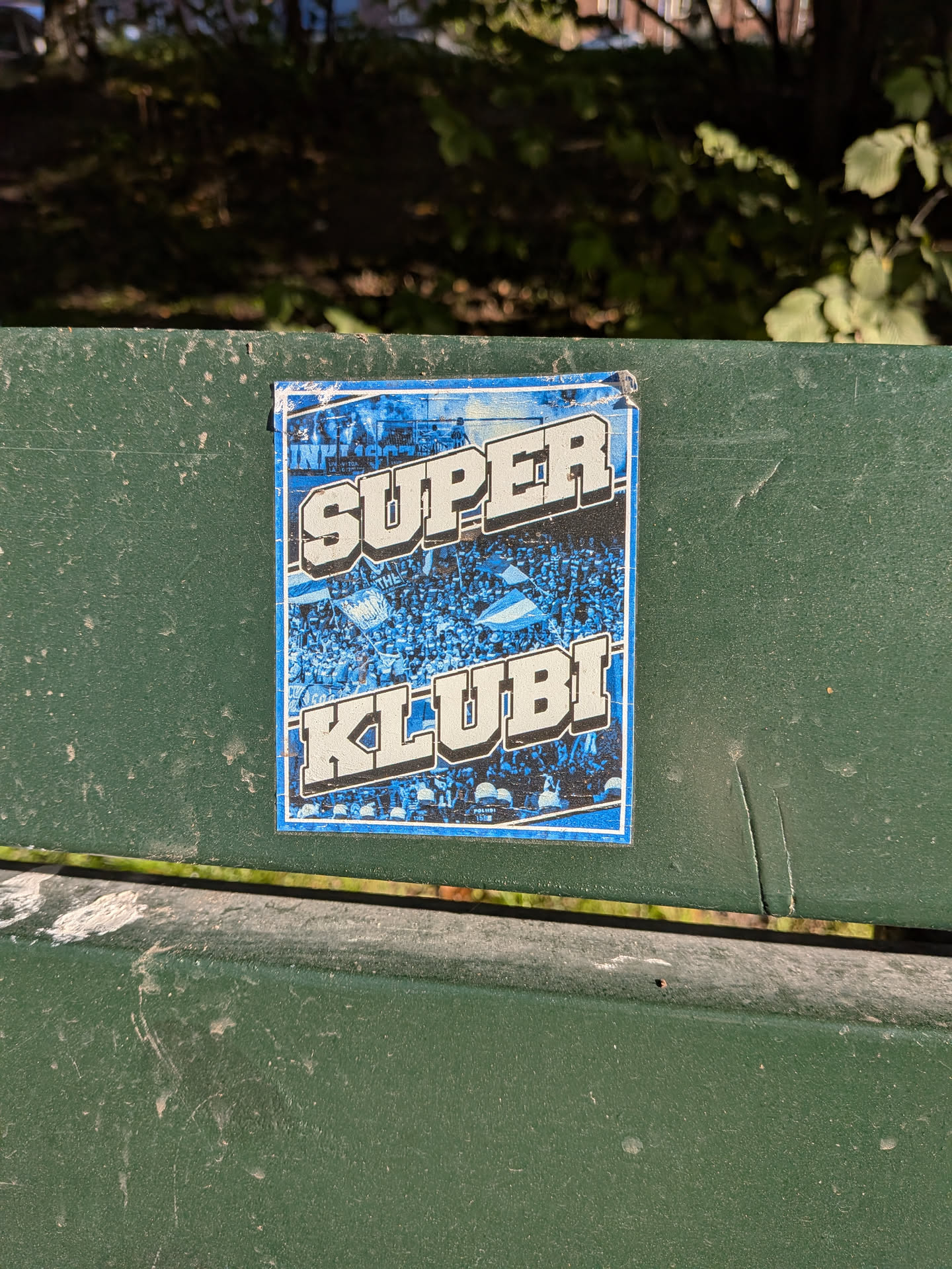 The sticker is predominantly blue and white, featuring a dense crowd of people with their arms raised, many holding flags. The word 'SUPER' is emblazoned in large, bold, white, 3D-effect letters across the top half of the sticker, with a blue background behind it. Below 'SUPER', the word 'KLUBL' is similarly presented in large, bold, white, 3D-effect letters. The background behind the crowd appears to be a stylized representation of a stadium or a large gathering, with a blue and white color scheme. The edges of the sticker show some wear and tear, indicating it has been applied for some time.