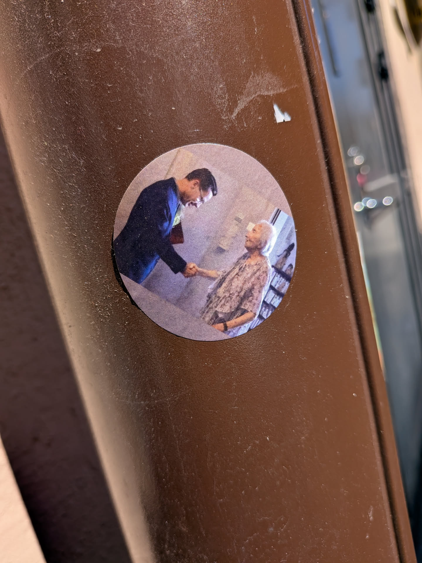A circular sticker featuring a photograph of a man in a dark suit jacket and glasses, with dark hair, shaking hands with an elderly woman. The man is positioned on the left, facing right, with his head slightly bowed. The woman, on the right, has white or grey hair and is wearing a light-colored, possibly patterned blouse, looking up at the man. The background is softly blurred, suggesting an indoor setting with faint details of shelves or furniture. The overall color scheme is muted, dominated by blues, greys, and light browns, with a slightly grainy texture on the sticker's surface.