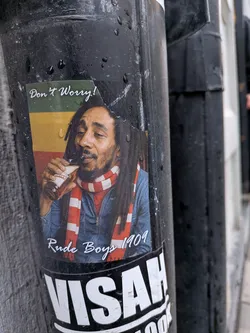 Street sticker The sticker features a portrait of Bob Marley, depicted from the chest up, with his characteristic dreadlocks and beard. He is wearing a blue denim jacket and a red and white striped scarf around his neck. He holds a brown bottle, partially labeled 'WILD TIGER', to his mouth, appearing to drink from it with a relaxed expression. The background behind him consists of horizontal stripes in green, gold (yellow), and red, reminiscent of the Rasta flag. Above his head, in the top left, is the white cursive text 'Don't Worry!'. Below his image, at the bottom center, is the white cursive text 'Rude Boys 1909'. The rectangular sticker is affixed to a dark pole and shows signs of wear and water droplets.