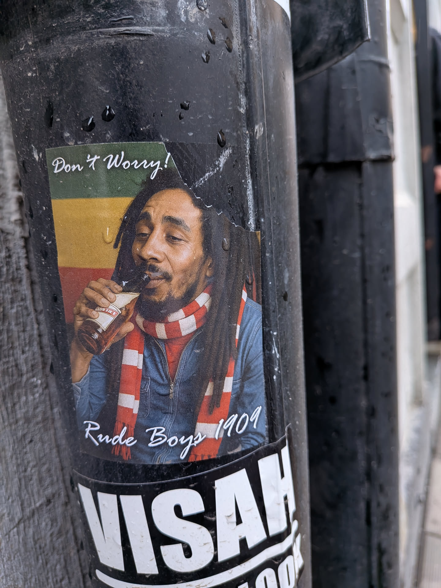The sticker features a portrait of Bob Marley, depicted from the chest up, with his characteristic dreadlocks and beard. He is wearing a blue denim jacket and a red and white striped scarf around his neck. He holds a brown bottle, partially labeled 'WILD TIGER', to his mouth, appearing to drink from it with a relaxed expression. The background behind him consists of horizontal stripes in green, gold (yellow), and red, reminiscent of the Rasta flag. Above his head, in the top left, is the white cursive text 'Don't Worry!'. Below his image, at the bottom center, is the white cursive text 'Rude Boys 1909'. The rectangular sticker is affixed to a dark pole and shows signs of wear and water droplets.