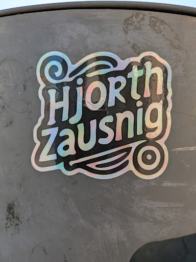 The sticker features a holographic effect, giving it a shimmering appearance. The text "Hjorth Zausnig" is prominently displayed in a stylized font. Below the text is a graphic of a stylized leaf design with a circle at the end, adding a whimsical touch.