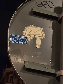 A beige sticker featuring a robot carrying an envelope. The robot has a simple design with circular arms and legs. The sticker has a slightly worn look, suggesting it has been affixed outdoors for some time. The sticker is adhered to a dark grey traffic sign, partially obscuring another sticker underneath.