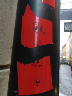 A sticker on a black pole. The sticker is primarily red with a distressed, torn look. There are black skull-like images printed on it. The style is abstract and dark.