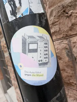 Street sticker The sticker is circular, featuring a central black and white illustration of an intricate, vintage electronic or scientific apparatus, complete with dials, switches, and a screen. This illustration is set against a fragmented, abstract background composed of torn-paper-like shapes in soft pastel shades of light blue, pale yellow, and light pink. A small, light beige circle with a dark blue center is positioned in the lower-left area of the background. Below the main illustration, black text reads "Das Auto fährt gegen die Wand.", with the word "gegen" in bold and "die Wand" highlighted in yellow. The sticker shows some signs of wear and tear, particularly around its edges and where it adheres to the pole.