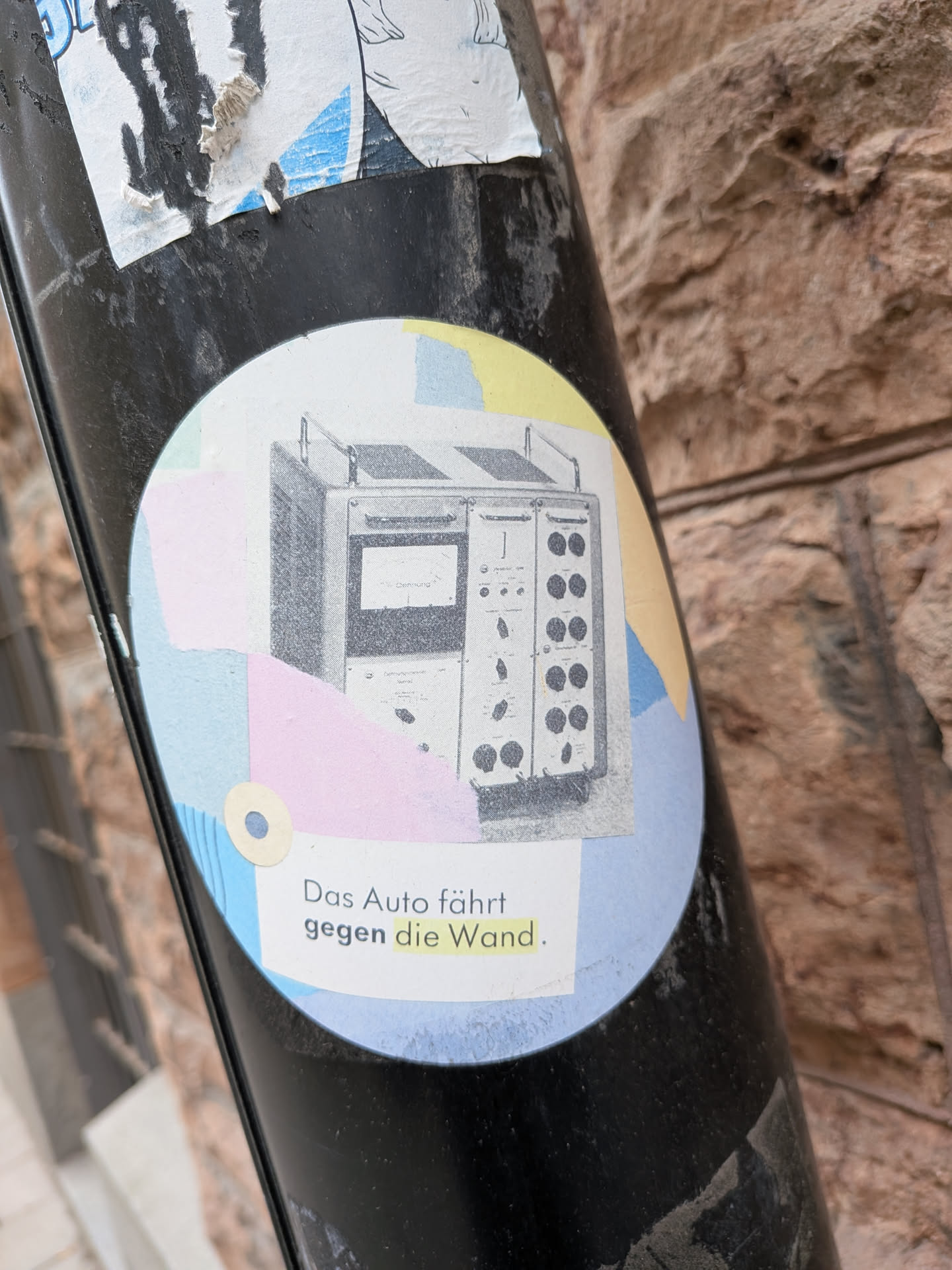The sticker is circular, featuring a central black and white illustration of an intricate, vintage electronic or scientific apparatus, complete with dials, switches, and a screen. This illustration is set against a fragmented, abstract background composed of torn-paper-like shapes in soft pastel shades of light blue, pale yellow, and light pink. A small, light beige circle with a dark blue center is positioned in the lower-left area of the background. Below the main illustration, black text reads "Das Auto fährt gegen die Wand.", with the word "gegen" in bold and "die Wand" highlighted in yellow. The sticker shows some signs of wear and tear, particularly around its edges and where it adheres to the pole.