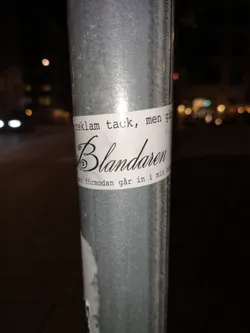 Street sticker A white rectangular sticker is affixed to a textured, galvanized metal pole at night. The sticker features black text arranged in three lines. The top line, 'reklam tack, men gå', and the bottom line, 'mot förmodan går in i min bo', are in a simple, serif-like font. The central and most prominent word, 'Blandaren', is rendered in an elaborate, elegant calligraphic script with a large, decorative initial 'B'. The sticker shows minor wear, with a slight curl at its bottom edge. The background is blurred, showing distant streetlights and vehicle lights.