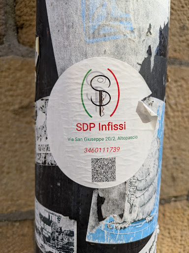 A round SDP Infissi sticker is pasted on a pole. The logo features a stylized key encircled by green and red lines, reminiscent of the Italian flag. Below the logo is the company name "SDP Infissi," followed by their address "Via San Giuseppe 20/2, Altopascio" and a phone number, "3460111739". A QR code is located at the bottom.