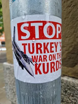 A sticker depicting a fighter jet and the text "STOP TURKEY'S WAR ON THE KURDS" in red and white. The sticker is affixed to a metal pole outdoors and shows water droplets.