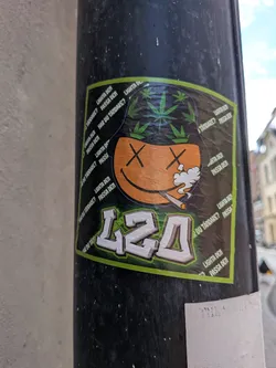 Street sticker A square sticker with a green border depicting an orange smiley face with 'X' eyes smoking a joint. The character wears a black bucket hat decorated with green cannabis leaves. Below is the stylized text '420' in white and green. The dark background features repeating Swedish phrases like 'LIGHTA DEN' and 'PASSA DEN'.