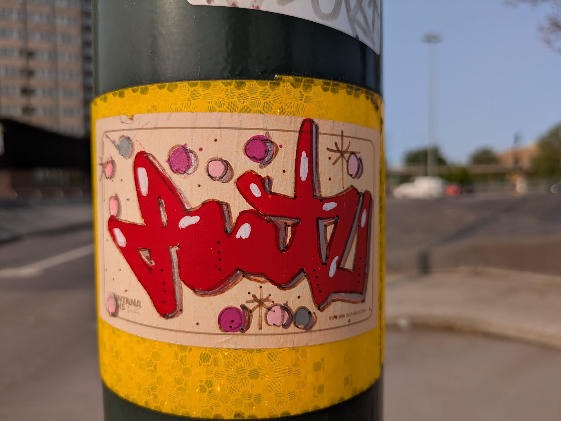 The sticker features the word "Antz" written in a bold, red graffiti style with white highlights. The background is a light tan color with small dots and circles in various shades of pink and gray, adding a playful and chaotic feel. The sticker has a cream colored border and the sticker is affixed to a pole covered in yellow reflective tape.