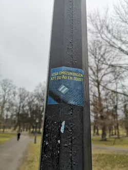 Street sticker The sticker is rectangular with slightly rounded corners, featuring a dark blue background with a subtle textured pattern, possibly resembling denim or a woven fabric. Prominently displayed in the upper left is yellow, uppercase text. Below the text, towards the center-right, is a light blue or white diamond-shaped insignia with two horizontal stripes inside, reminiscent of a military rank or badge. The sticker appears slightly worn, with some edges lifting from the dark grey lamppost it's affixed to.