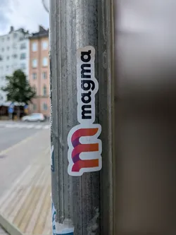 Street sticker A die-cut sticker on a metal pole displaying the word 'magma' in black lowercase text vertically above a stylized 'm' logo with a purple-to-orange gradient ribbon design.