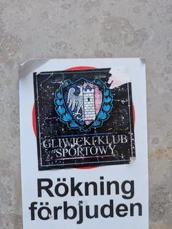 Street sticker A square, weathered black sticker showing the emblem of Gliwicki Klub Sportowy. The crest features a split shield with a white eagle and a stone tower, surrounded by a blue wreath. Below, the text 'GLIWICKI KLUB SPORTOWY' is written in white. The sticker is scratched and peeling at the top right.
