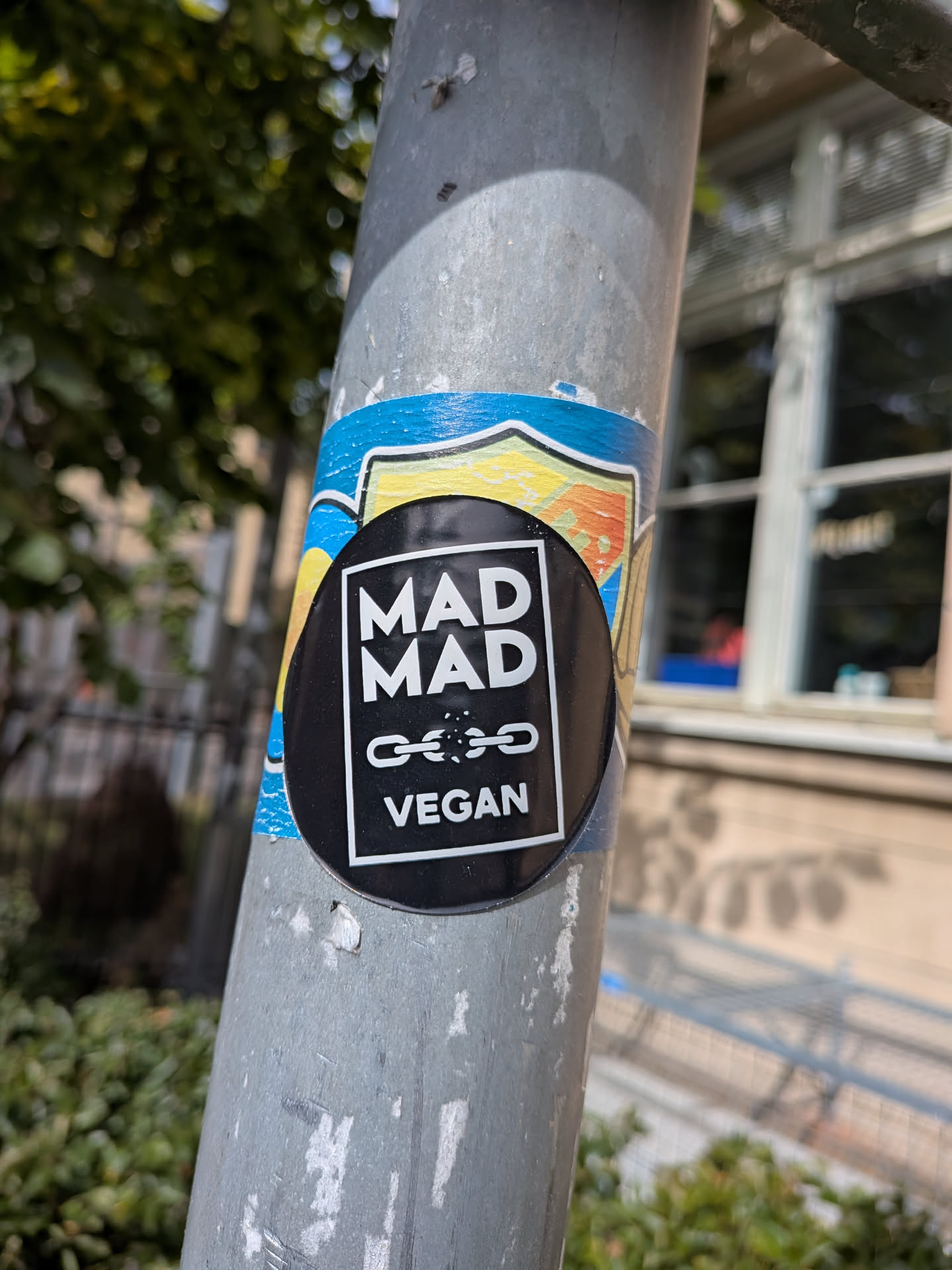 A circular black sticker with a white rectangular border. Inside, the text 'MAD MAD' is stacked above a broken chain graphic, with 'VEGAN' at the bottom. It is pasted on a metal pole.