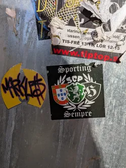 Street sticker The central sticker is a square-shaped, predominantly black sticker. At the top, in a white gothic-style font, is the word "Sporting". Below this, the letters "SCP" (likely an acronym for Sporting Clube de Portugal) are displayed in white, flanked by white laurel wreaths. Further down, two shield-shaped crests are featured: the left one depicts the Portuguese national shield (red border with castles, white background with blue shields), and the right one is green with a white rampant lion, the emblem of Sporting CP. To the right of these crests is a stylized white 'S' within another shield outline. At the bottom, in the same gothic-style font, is the word "Sempre" (Portuguese for 'Always'). The sticker has a slightly worn top edge.