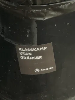 Street sticker A square-shaped sticker with a dark brown-grey background and white text. The main text, 'KLASSKAMP UTAN GRÄNSER' (Class Struggle Without Borders), is prominently displayed in bold, sans-serif capital letters, stacked in three lines. Below this, in smaller white text, is 'Allt åt alla' (Everything for everyone). To the left of the smaller text is a white circular symbol, commonly recognized as an anarchist 'A' within a circle. The sticker shows signs of wear, with some scratches and a slightly peeling corner, indicating it's affixed to a textured, dark surface.
