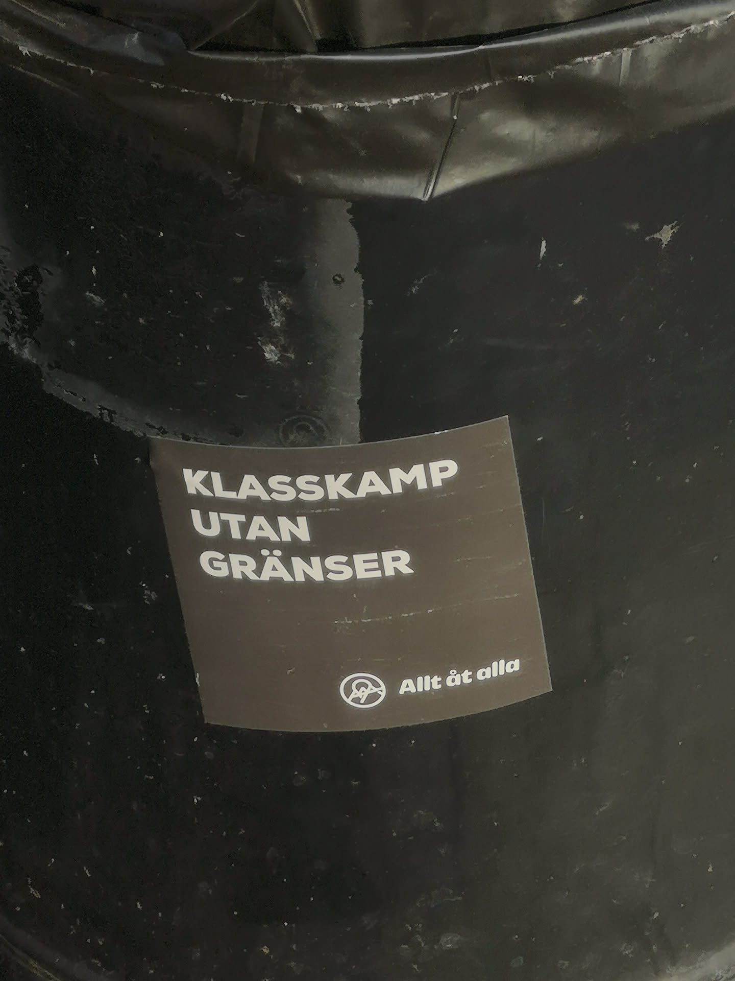 A square-shaped sticker with a dark brown-grey background and white text. The main text, 'KLASSKAMP UTAN GRÄNSER' (Class Struggle Without Borders), is prominently displayed in bold, sans-serif capital letters, stacked in three lines. Below this, in smaller white text, is 'Allt åt alla' (Everything for everyone). To the left of the smaller text is a white circular symbol, commonly recognized as an anarchist 'A' within a circle. The sticker shows signs of wear, with some scratches and a slightly peeling corner, indicating it's affixed to a textured, dark surface.