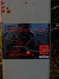 Street sticker A dark, glossy sticker is adhered to a weathered, rust-stained white pole outdoors. The sticker features a striking design with red text and graphics against a black background that appears to have an undulating, liquid-like texture. At the top, 'New music from:' is written in white, followed by 'A BRAND NEW STAGE' in bold, red uppercase letters. Below this, an abstract outline of a profile or face, rendered in red, merges with the textured background. Towards the bottom left, the word 'Sensuality' is written in a flowing, stylized red script, accompanied by a subtle red glow. On the right side, a prominent red QR code with a green Spotify logo at its center is visible. Beneath the QR code, a small grey rectangular button contains the text 'SKANNA MIG' in white.